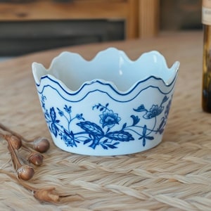 May include: A white ceramic bowl with a scalloped rim and blue floral patterns. The bowl has a wavy blue border and is set on a woven surface. The floral design includes detailed leaves and flowers.