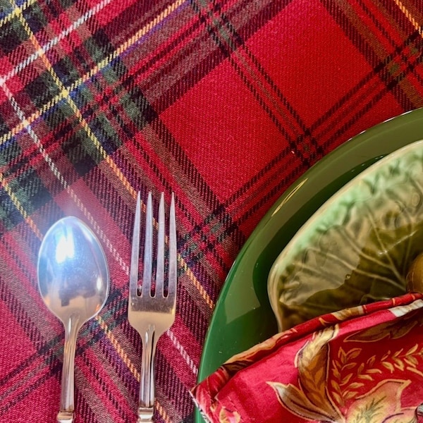 May include: A stainless steel spoon and fork with red handles placed on a red, green, and yellow plaid tablecloth. A dark green plate sits partially visible with a folded red napkin featuring a floral pattern resting on top of the plate.
