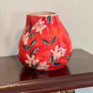 May include: A red ceramic vase with a bulbous shape. The vase is decorated with white floral patterns and green leaves. The interior of the vase is a light beige color. The vase is sitting on a dark brown surface.