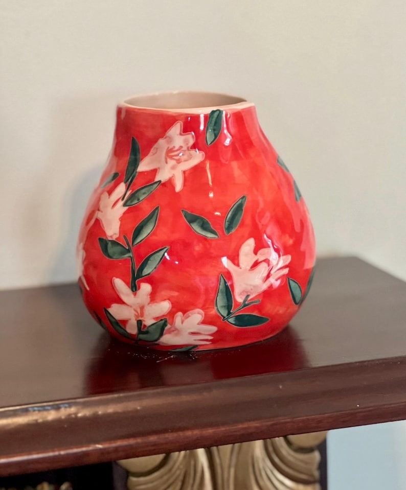 May include: A red ceramic vase with a bulbous shape. The vase is decorated with white floral patterns and green leaves. The interior of the vase is a light beige color. The vase is sitting on a dark brown surface.