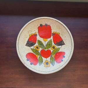 May include: A round, off-white ceramic plate with a hand-painted floral design. The design features red tulips, birds, and hearts, with green leaves and yellow flowers. The plate has a scalloped edge.