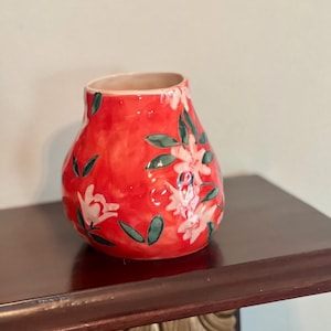 May include: A red ceramic vase with a floral design. The vase has a rounded shape and a wide opening. The flowers are white with green leaves. The vase is sitting on a dark brown surface.