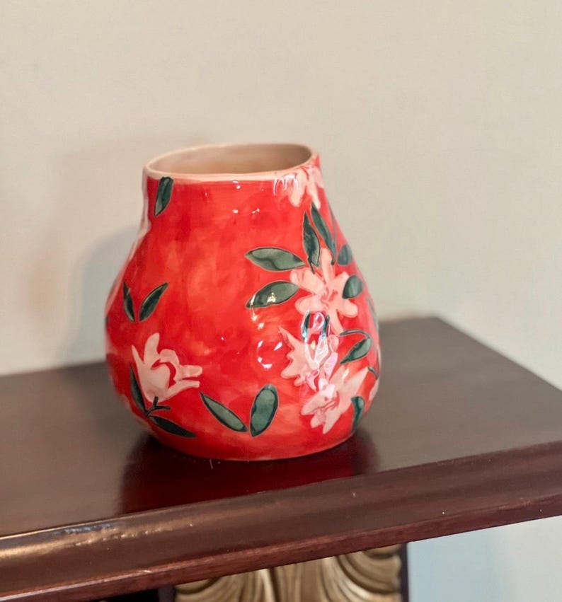 May include: A red ceramic vase with a floral design. The vase has a rounded shape and a wide opening. The flowers are white with green leaves. The vase is sitting on a dark brown surface.