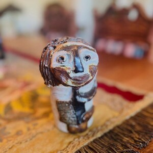 May include: A small, hand-sculpted ceramic figurine featuring a stylized human face with dark brown hair, deep-set eyes, and a prominent nose. The figure has a glossy, multi-toned glaze in shades of cream, dark brown, and black, with a textured surface.