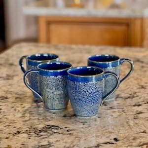 May include: Four blue ceramic mugs with textured exteriors and dark blue interiors and rims. The mugs have curved handles and are arranged on a speckled countertop. The mugs are a matching set.