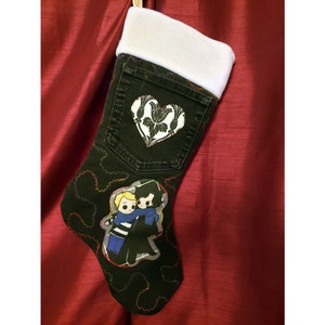 May include: A black denim Christmas stocking with a white fleece cuff. The stocking features a white heart-shaped patch with a floral design and a cartoon illustration of two figures hugging. Colorful stitching outlines the design.