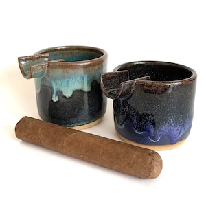 May include: Two ceramic ashtrays with a unique design. The ashtrays are dark brown with blue and green glaze. One ashtray has a blue glaze and the other has a green glaze. A brown cigar is in the foreground.
