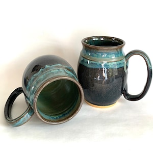 May include: Two handmade ceramic mugs. The mugs are dark blue with teal and brown accents. One mug is upright, and the other is on its side. The interior of the mugs is green. The mugs have large handles.
