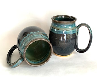 May include: Two handmade ceramic mugs. The mugs are dark blue with teal and brown accents. One mug is upright, and the other is on its side. The interior of the mugs is green. The mugs have large handles.