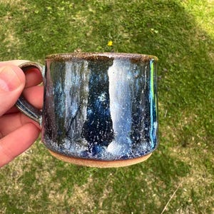 May include: A ceramic mug featuring a glossy, mottled glaze in shades of deep navy blue, black, and white, with a textured, dark brown rim and a natural, unglazed terracotta-colored base. The mug includes a curved handle on the left side.