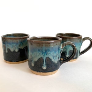 May include: Three handmade ceramic mugs with a unique glaze. The mugs feature a dark brown rim that transitions into shades of blue and black, with a dripped glaze effect. The mugs have a natural, unglazed base and a curved handle.