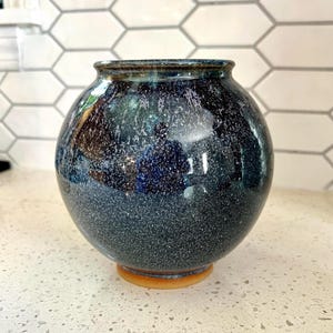 May include: A round, speckled ceramic vase with a dark blue and teal glaze. The vase has a wide body and a narrow opening, sitting on a small, light brown base. The glaze has a glossy finish, reflecting light.