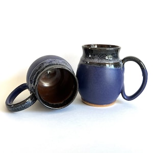 May include: Two handmade ceramic mugs with a dark blue glaze and a black rim. The mugs have a speckled glaze and are slightly rounded.