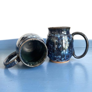 May include: Two handmade ceramic mugs with a dark blue glaze and speckled pattern. One mug is upright, the other is on its side. Both have large handles and a natural clay base. The mugs are on a blue surface.