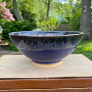 May include: A ceramic bowl with a wide, circular opening and a tapered base. The exterior features a deep blue glaze at the bottom that transitions into a dark, mottled black and grey finish toward the rim. The base is unglazed, showing a light tan color.