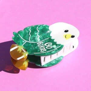 May include: A green and white eagle-shaped hair clip with the words "GO BIRDS" in white. The clip features a football accent and is set against a pink background. The eagle has a white head, black eyes, and a yellow beak.