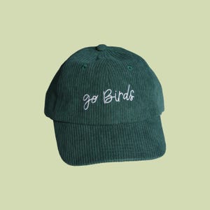 Könnte beinhalten: Dunkelgrüne Cord-Baseballkappe mit dem gestickten Text "go Birds" in weißer Schrift. Die Kappe hat einen gebogenen Schirm und einen Knopf oben. Ein Hut im klassischen Stil, geeignet für Freizeitkleidung.
