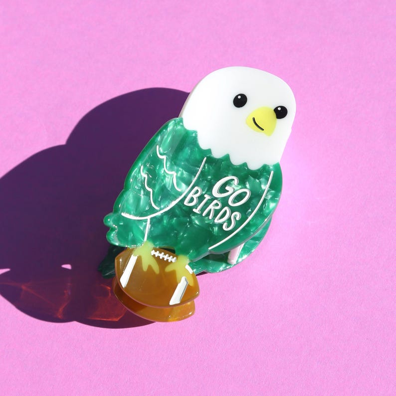 May include: A decorative eagle-shaped pin with a white head, green body, and a brown football. The pin has the words "GO BIRDS" in white. The background is a solid pink colour.
