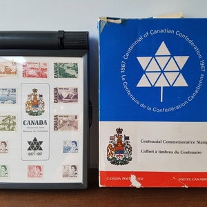 May include: A black box containing a collection of Canadian stamps commemorating the 100th anniversary of Canadian Confederation in 1967. The box is closed and has a clear plastic window showing the stamps. The box is sitting on a wooden surface. The box is labelled "Centennial Commemorative Stamp Box" and "Coffret à timbres du Centenaire".