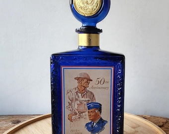 May include: A rectangular cobalt blue glass decanter with a matching round stopper featuring a gold-colored emblem. The front label depicts two figures in uniform with the text 50th Anniversary and 1919-1969. The bottle has a gold-colored neck band.