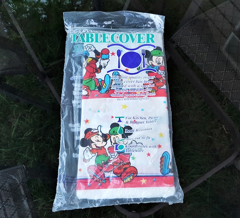以下が含まれることがあります： 透明なビニール袋に入ったプラスチック製のテーブルクロス。ミッキーとミニーマウスが冬の衣装を着ています。テーブルクロスには「TABLECOVER」、「For Kitchen, Picnic & Banquet Tables」、「Leak Resistant」と書かれています。