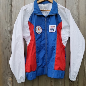 May include: A blue, red, and white windbreaker jacket with a zipper closure. The jacket has a white patch with the text "Team BC" and a red patch with a flame design.