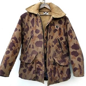 Puede incluir: Chaqueta de camuflaje marrón y beige con cuello de pana. La chaqueta tiene cierre de cremallera, dos bolsillos delanteros y un parche circular en el pecho. La chaqueta está colgada en una percha de madera.