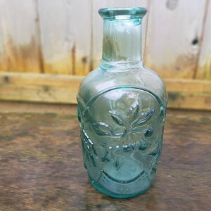Peut inclure: Une bouteille en verre transparente de couleur vert d'eau, avec un corps arrondi et un col &eacute;troit. La bouteille pr&eacute;sente des motifs floraux en relief et mesure environ 20 cm de haut. La bouteille est pos&eacute;e sur une surface en bois.