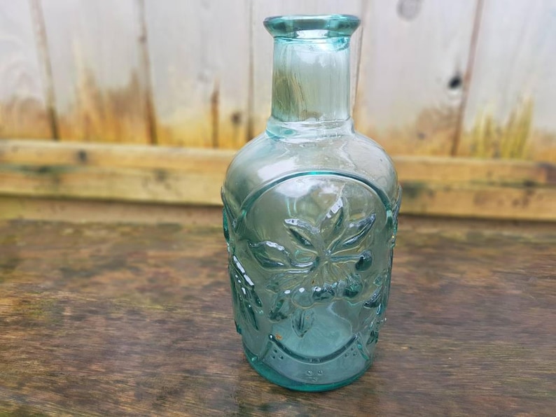 Peut inclure: Une bouteille en verre transparente de couleur vert d'eau, avec un corps arrondi et un col &eacute;troit. La bouteille pr&eacute;sente des motifs floraux en relief et mesure environ 20 cm de haut. La bouteille est pos&eacute;e sur une surface en bois.