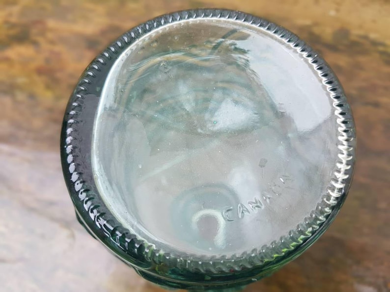 Peut inclure: Base d'une bouteille en verre clair, teint&eacute; de vert, avec un bord textur&eacute;. Le mot "CANADA" est estamp&eacute; au fond. La conception de la bouteille sugg&egrave;re qu'elle peut &ecirc;tre utilis&eacute;e pour des boissons ou &agrave; des fins d&eacute;coratives.