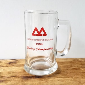 Puede incluir: Jarra de cerveza de vidrio transparente con asa. La jarra tiene texto rojo que dice "Alberni Pacific Division 1994 Grading Championship" y un logotipo rojo. La jarra está sobre una superficie de madera.