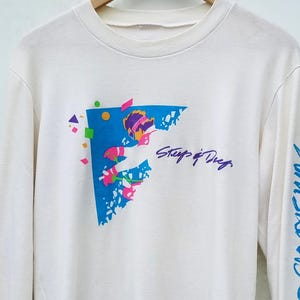 Puede incluir: Camiseta de manga larga color crema con un diseño gráfico colorido. El diseño presenta una forma azul con el texto "Steep & Deep" y "Whistler BC" en escritura azul en la manga. El gráfico incluye acentos morados, rosas y verdes.