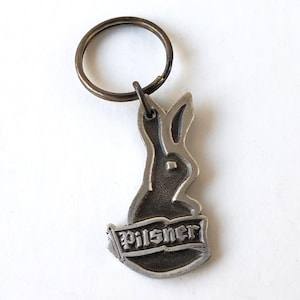 May include: A silver-coloured metal keyring featuring a rabbit design with the word "Pilsner" on a banner. The keyring has a circular ring for attaching keys.