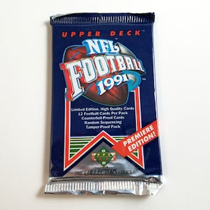 Könnte beinhalten: Ein blaues und rotes Upper Deck NFL Football 1991 Sammelkarten-Paket. Das Paket zeigt einen Fußball mit dem Text "NFL Football 1991" und die Worte "Premiere Edition!" in einem roten Banner.