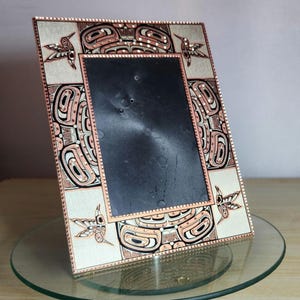 Puede incluir: Un marco de fotos rectangular con interior negro y borde decorativo. El borde presenta un fondo crema con diseños geométricos salmón, negros y blancos e ilustraciones de colibríes. El marco se muestra sobre una superficie de vidrio.