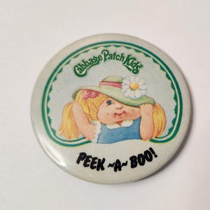 Puede incluir: Un botón redondo con un borde verde y un fondo blanco. El botón presenta una muñeca Cabbage Patch Kid con cabello rubio que lleva un sombrero verde con una flor blanca. El texto "Cabbage Patch Kids" está escrito en un círculo verde sobre la muñeca. El texto "Peek-a-Boo!" está escrito debajo de la muñeca.
