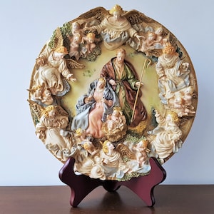 May include: A ceramic plate with a raised relief design depicting the Nativity scene. The plate features a central image of Mary, Joseph, and baby Jesus surrounded by a ring of angels. The plate is decorated with gold accents and has a brown wooden stand.