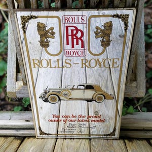 Könnte beinhalten: Vintage Rolls-Royce-Schild mit einer Illustration eines cremefarbenen und braunen Autos. Das Schild zeigt das Rolls-Royce-Logo, Text und ein Löwenemblem. Der Text lautet: "Sie können der stolze Besitzer unseres neuesten Modells sein".