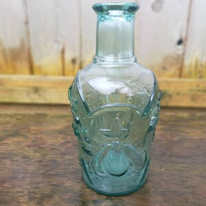 Peut inclure: Une bouteille en verre transparente de couleur vert-bleu avec un motif floral d&eacute;coratif. La bouteille a un long col et une base arrondie. La bouteille mesure environ 20 cm de haut.