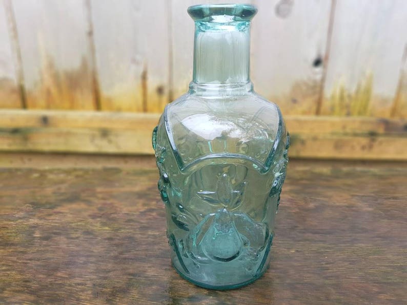Peut inclure: Une bouteille en verre transparente de couleur vert-bleu avec un motif floral d&eacute;coratif. La bouteille a un long col et une base arrondie. La bouteille mesure environ 20 cm de haut.