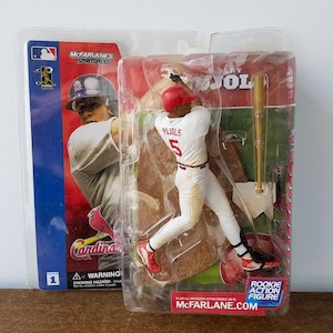 May include: A red and white baseball action figure of Albert Pujols, a former St. Louis Cardinals player, holding a bat. The figure is posed in a batting stance. The packaging includes the McFarlane Sports Picks logo and the words "Rookie Action Figure".