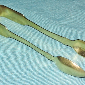 Puede incluir: Un par de pinzas de azúcar antiguas de plata, con un diseño en forma de U y extremos en forma de cuchara. Las pinzas tienen una textura acanalada en los mangos, sobre un fondo azul claro.