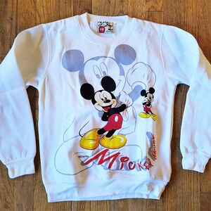 May include: White sweatshirt with a Mickey Mouse graphic print. The print features Mickey Mouse in a red and yellow outfit, with the text "Mickey Mouse" printed below.