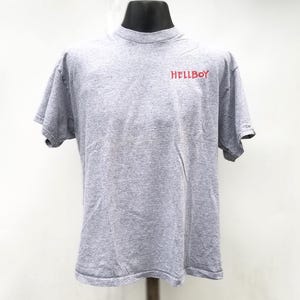 Puede incluir: Una camiseta gris jaspeada con la palabra "HELLBOY" impresa en rojo en el pecho. La camiseta tiene mangas cortas y cuello redondo. La prenda se exhibe en un maniquí negro.