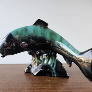 May include: A ceramic fish figurine with a black and green glaze. The fish is leaping out of the water and has a detailed fin and tail.