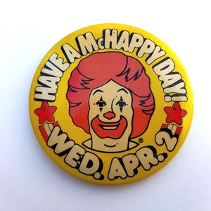 May include: A yellow button with the text "HAVE A McHAPPY DAY!" and "WED. APR. 2" encircling a cartoon clown face with red hair and a red nose. Red star and stripe graphics are on the button.
