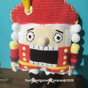 May include: A crocheted nutcracker doll in red, white, black, and yellow. The doll features a red hat with a yellow feather, a white face, and a black mouth. It wears a red jacket with yellow trim and black shoes.