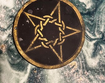 Puede incluir: Un medallón circular con una base de color marrón oscuro y un diseño de pentagrama dorado en el centro. El pentagrama está formado por un intrincado patrón de nudos entrelazados. El borde exterior del medallón tiene un acabado dorado.