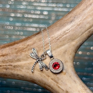 May include: A silver necklace featuring two pendants: a dragonfly and a pendant with a red gemstone. The dragonfly pendant is detailed with intricate wing designs. The gemstone pendant is round and set in a silver casing.