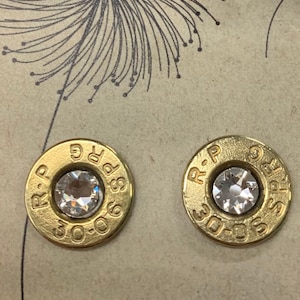 May include: A pair of gold-colored bullet casing earrings with clear rhinestones in the center. The casings are marked with "R-P 30-06" and "R-P 30-06".
